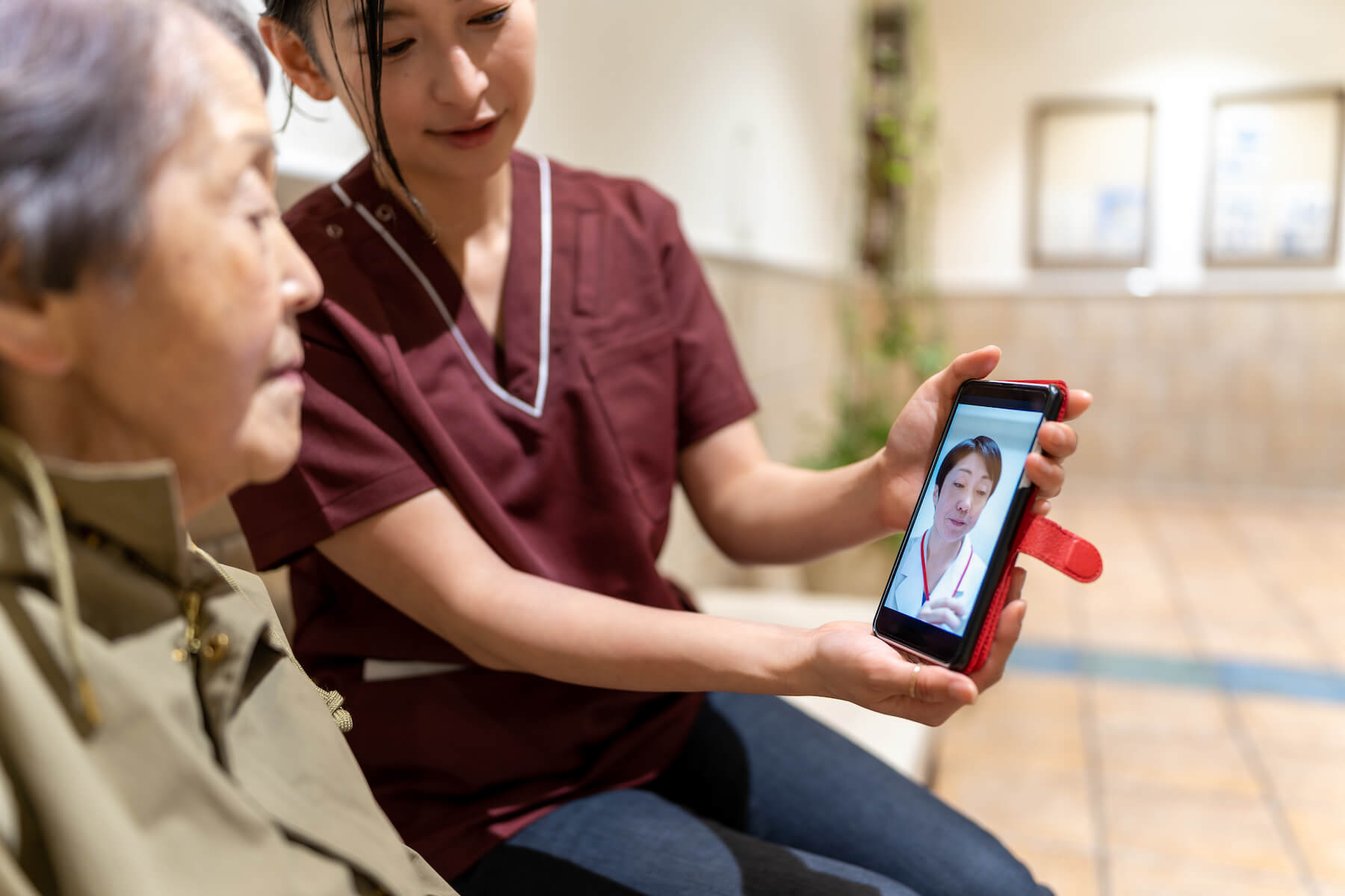 An Asian elder adult attends telehealth appointment by smartphone. A younger Asian woman family or staff members holds the mobile phone.