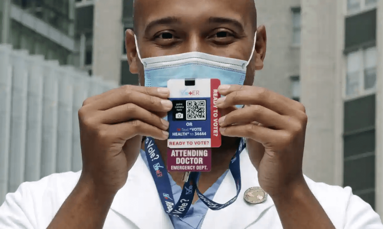 A light-skinned Black physician wearing a surgical mask holds up a voter information badge he wears on his ID