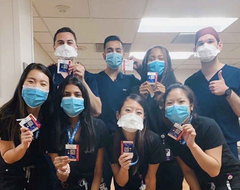 A racially diverse group of health care professionals in blue scrubs and surgical masks hold up voter registration badges