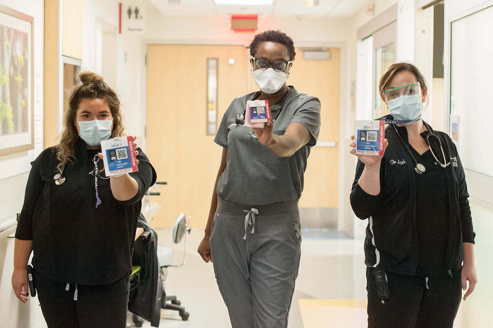 Three health care workers wearing scrubs in hospital hallway, each holding a a blue and red ID badge with a QR code in center and side label that reads, “Ready to Vote?”