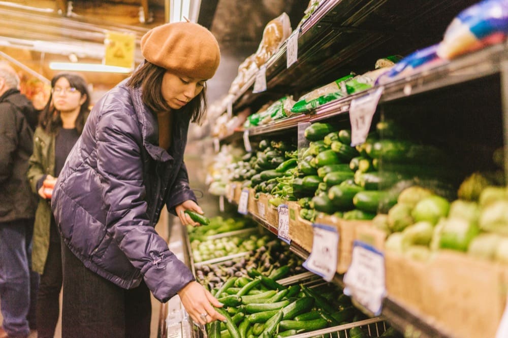 Asian woman in grocery store vegetable aisle reaching for cucumbers