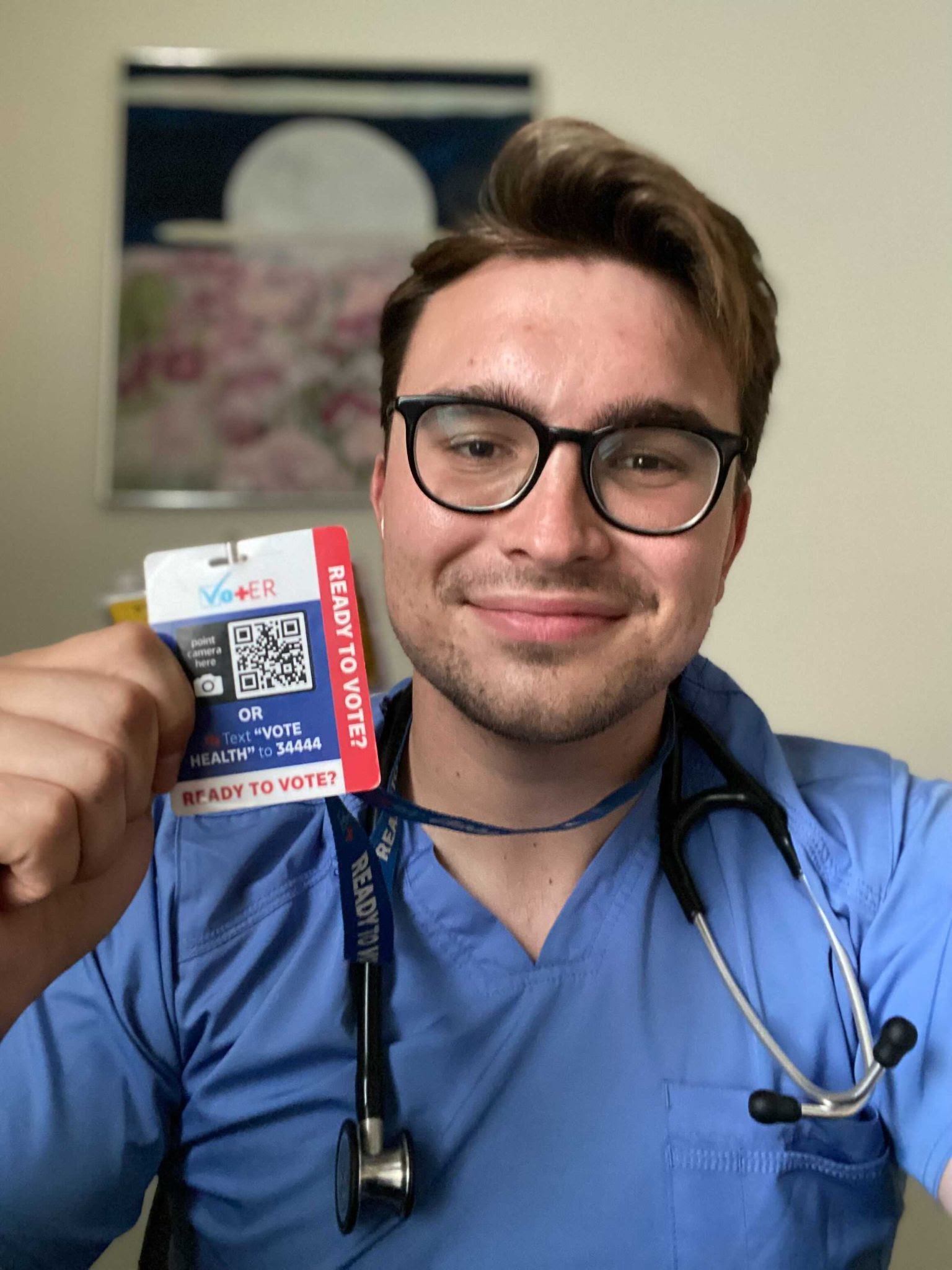 Young man in scrubs with stethescope around his neck; he holds a blue and red ID badge with a QR code in center and side label that reads, “Ready to Vote?”