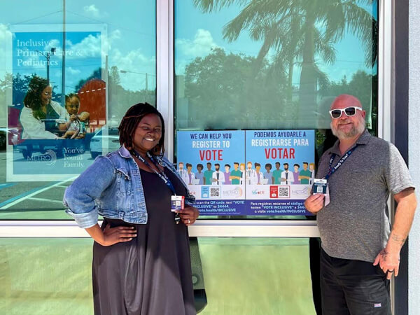 Two health care workers at stand in front of the entrance to a community health center, smiling with voter registration posterse with posters