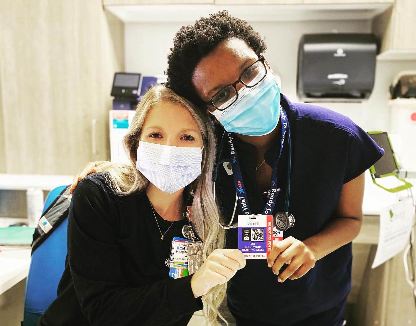 Two pediatric medical residents, one white and one Black, both masked, and posing with a a blue and red ID badge with a QR code in center and side label that reads, “Ready to Vote?”