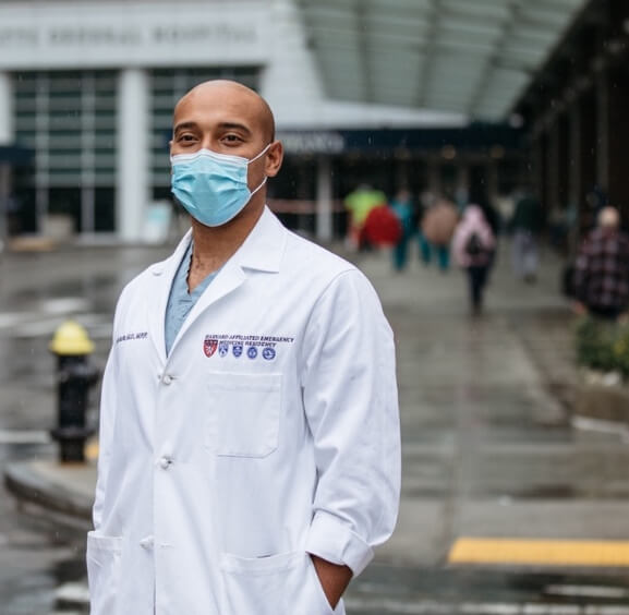 Physician in white coat and mask stands outside hospital